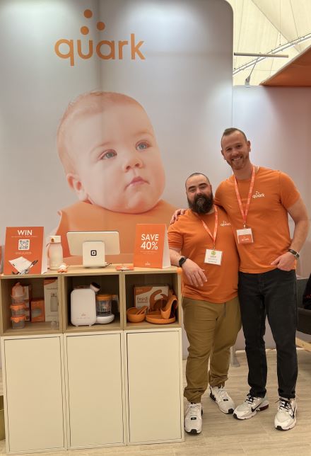 무역박람회 부스에서 제품 옆에 서 있는 Quark 공동창업자 Senez와 Gurinskas. 로고가 있는 뒷면 아기 사진.